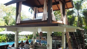 An outdoor bar area with a pool in the background, wooden furniture, and a covered seating area. - Lima Bella Resort (Rayong)