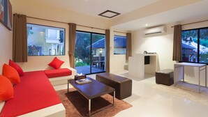 A modern living room with a sofa, ottoman, and coffee table. There are large windows with curtains, an air conditioning unit, and a view of a house outside. - Lima Bella Resort (Rayong)