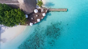 An aerial view of a wooden deck with lounge chairs and umbrellas, overlooking a turquoise sea and a sandy beach.