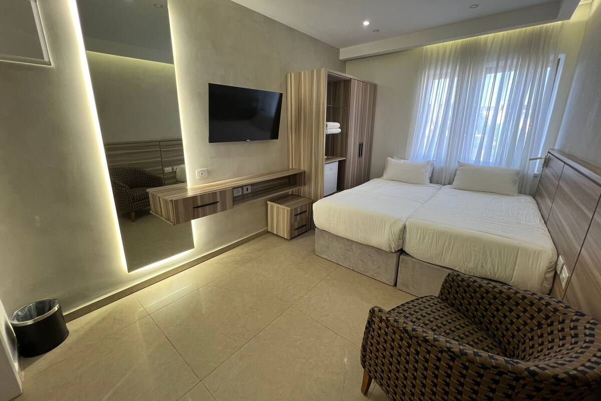 A modern hotel room with a large bed, a TV mounted on the wall, a seating area with a chair, and a wardrobe.