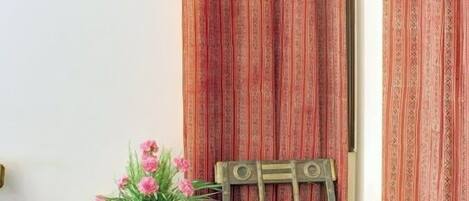 A corner room with a wooden chair, a small table with a vase of flowers, and red patterned curtains.