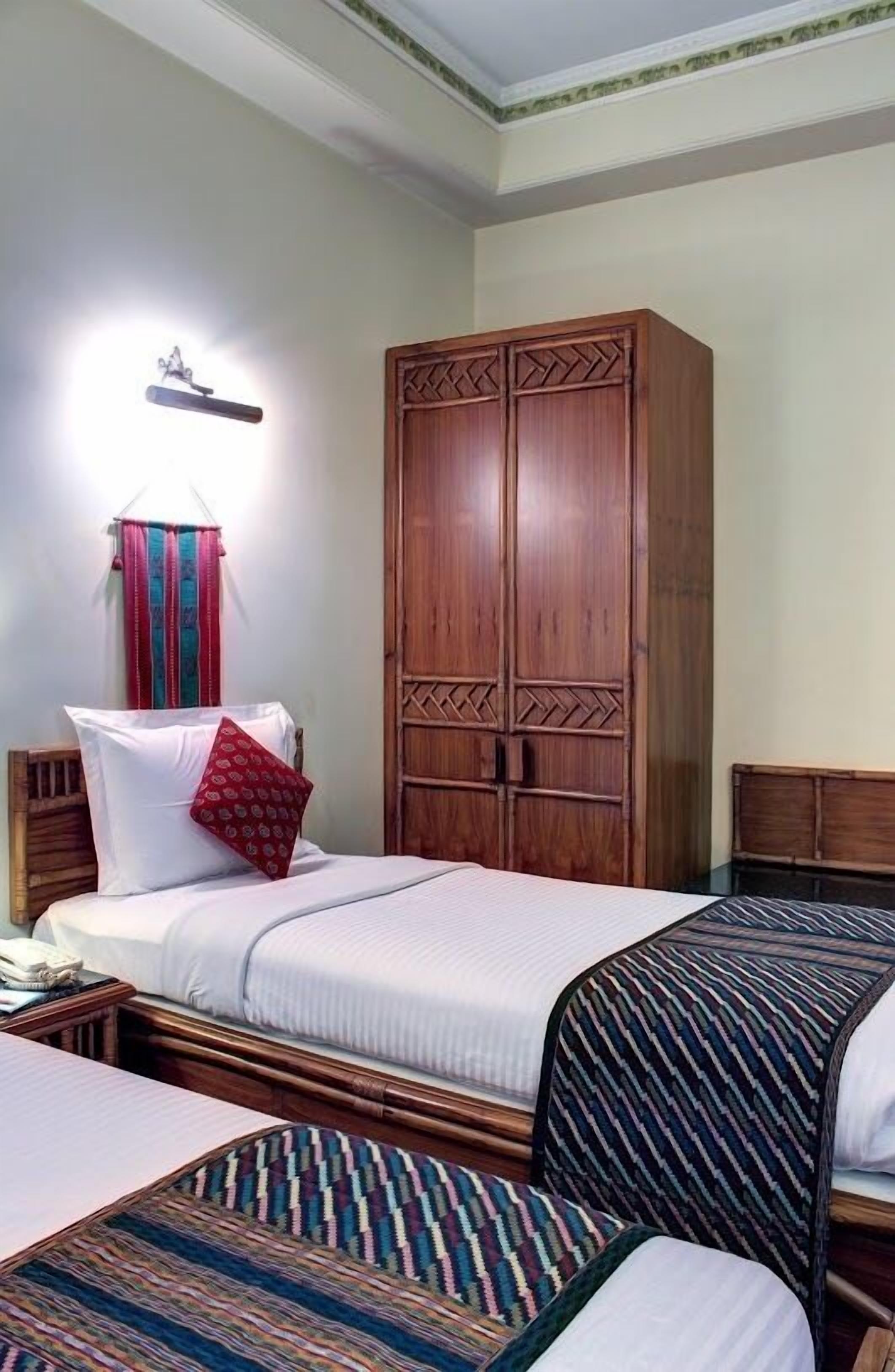 A hotel room with a large wooden wardrobe, a bed with white bedding, a colorful striped chair, and a small table with a telephone.