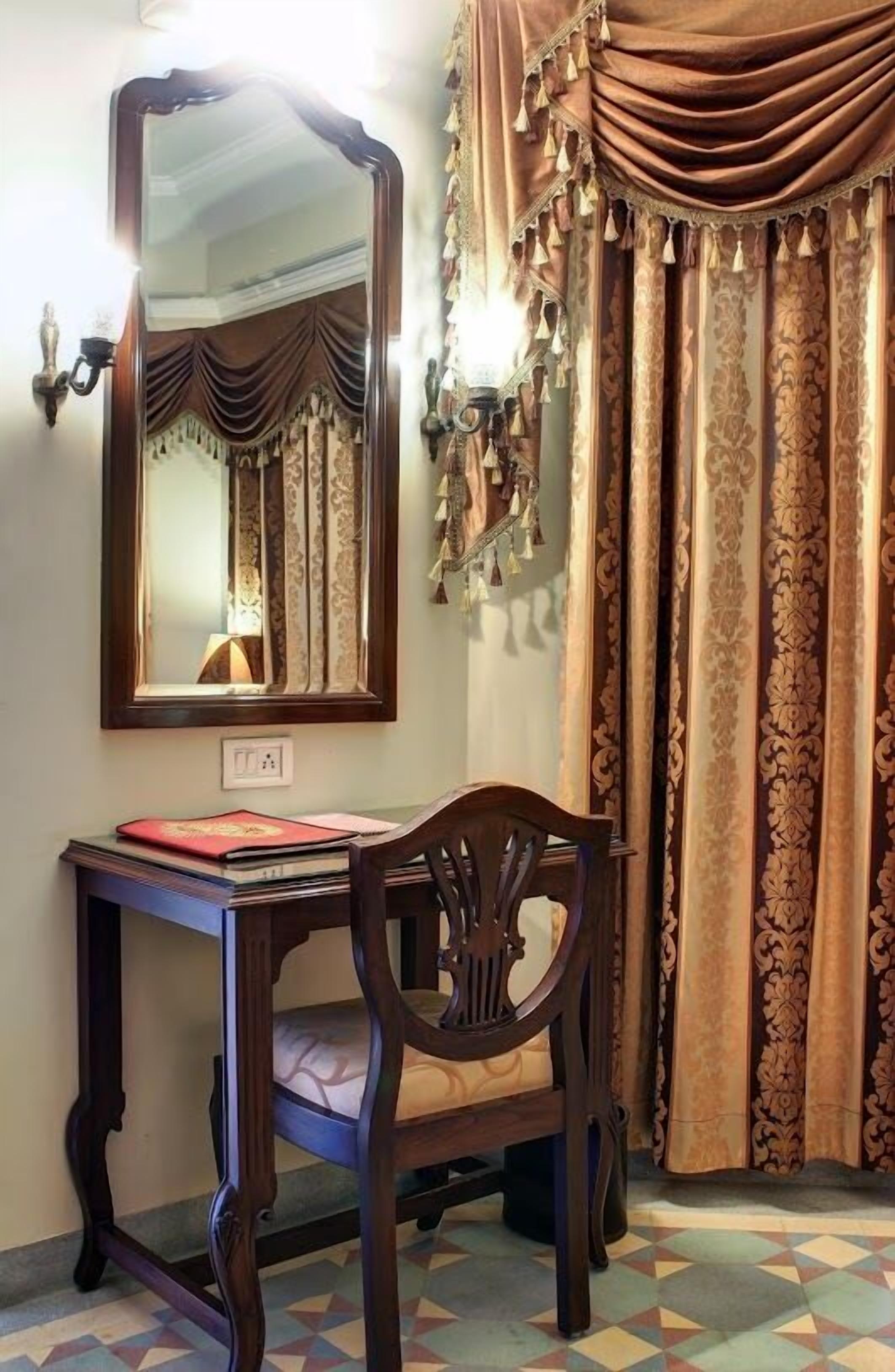 A room with a wooden desk, a mirror, and a chair. There is a lamp on the wall and curtains with tassels.