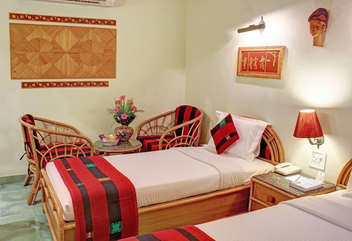 A hotel room with two beds, a small table with a vase of flowers, a chair, a nightstand with a lamp, and decorative wall art.