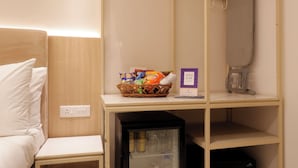 A compact hotel room with a bed, a small bedside table, a mini fridge, a shelf with snacks, a mini bar sign, and a hanging iron. - Hotel Varuna (Varanasi)