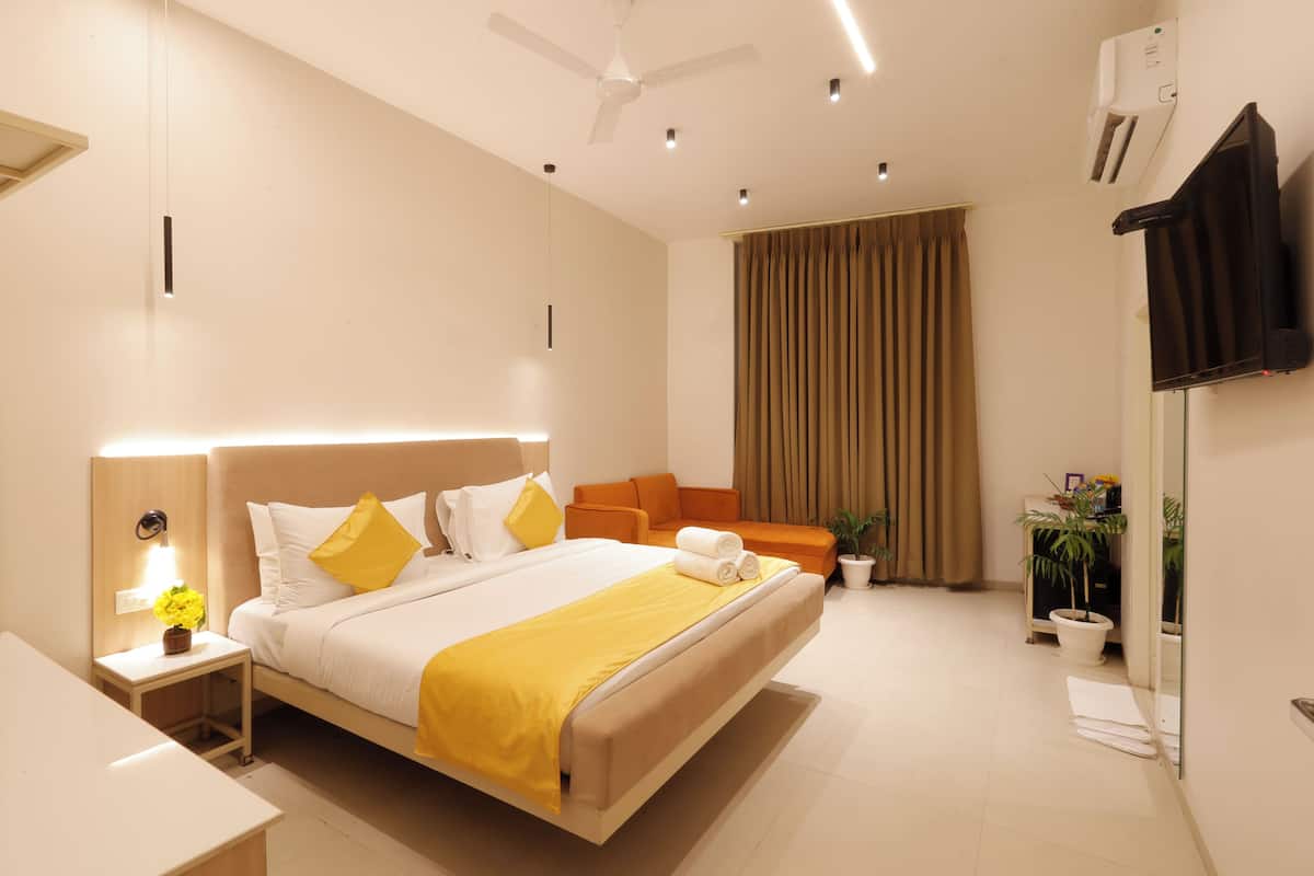 A modern hotel room with a large bed, a bedside table with a lamp, a small table with a vase, a sofa, a potted plant, and a flat-screen TV mounted on the wall.