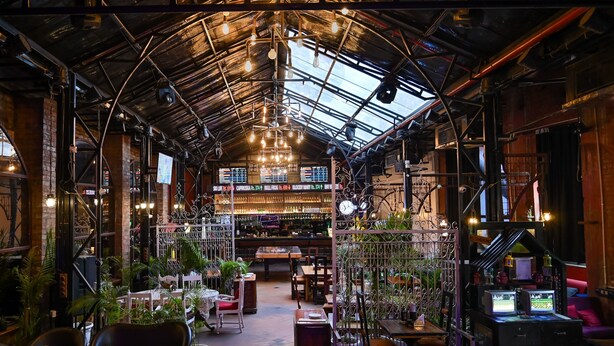 An industrial-style interior with exposed brick walls, metal beams, and a high ceiling with hanging lights. There are tables and chairs arranged for dining, and a bar area with a menu display. - Hotel Varuna (Varanasi)