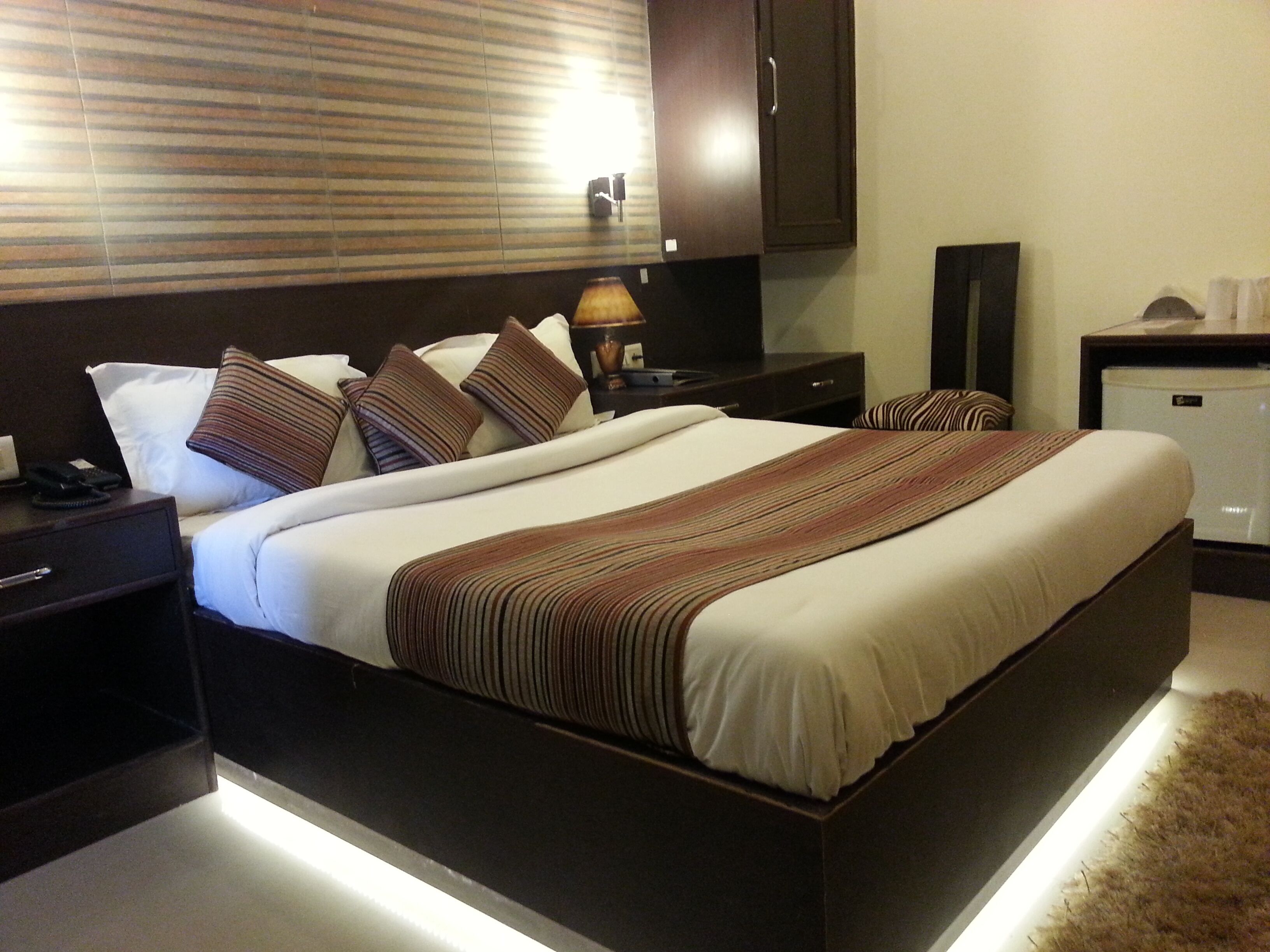 A hotel room with a large bed, dark wooden headboard, bedside tables with lamps, a nightstand, a small refrigerator, and a window with blinds.