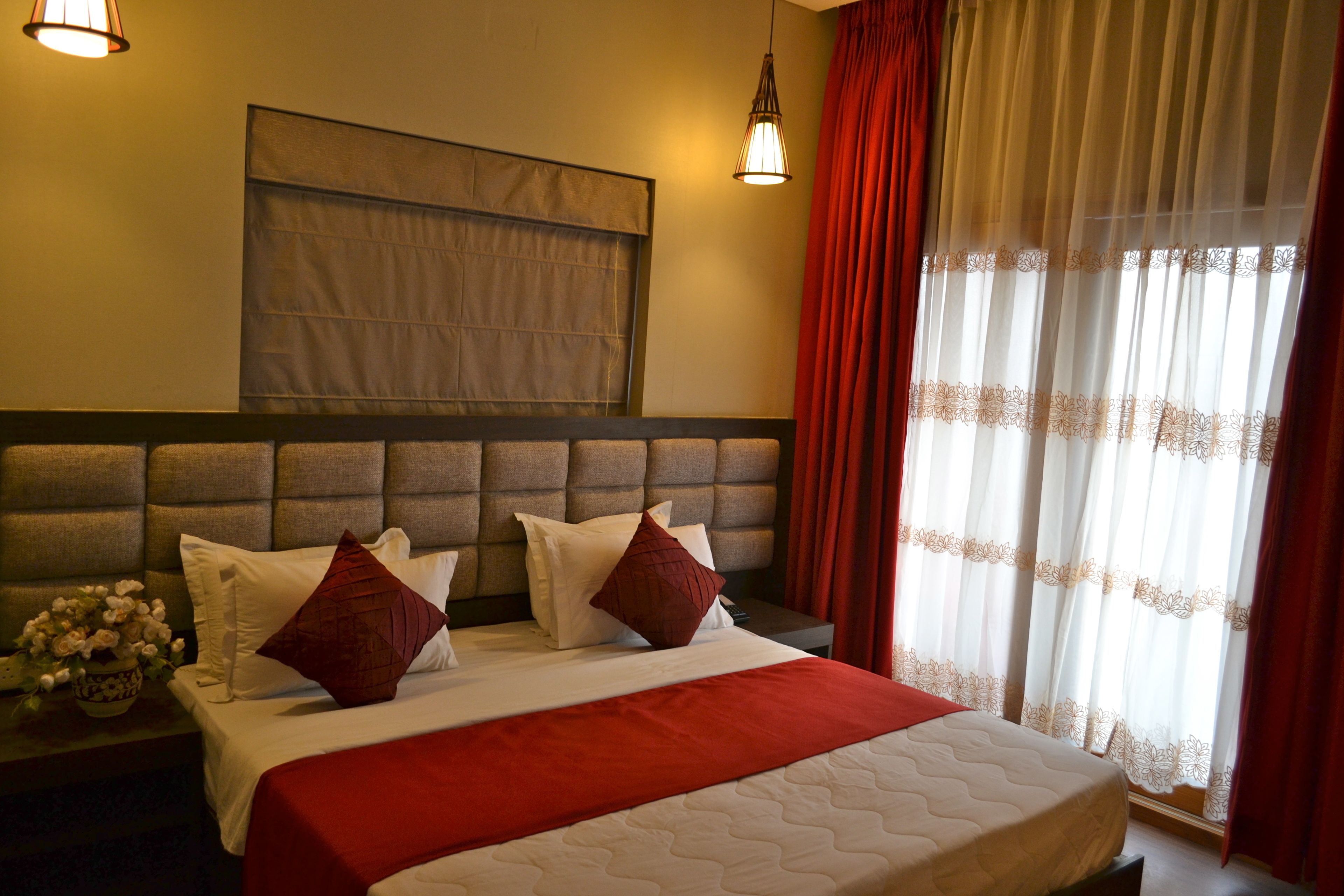 A hotel room with a large bed, a headboard, a bedside table with a vase of flowers, and a window with red curtains.