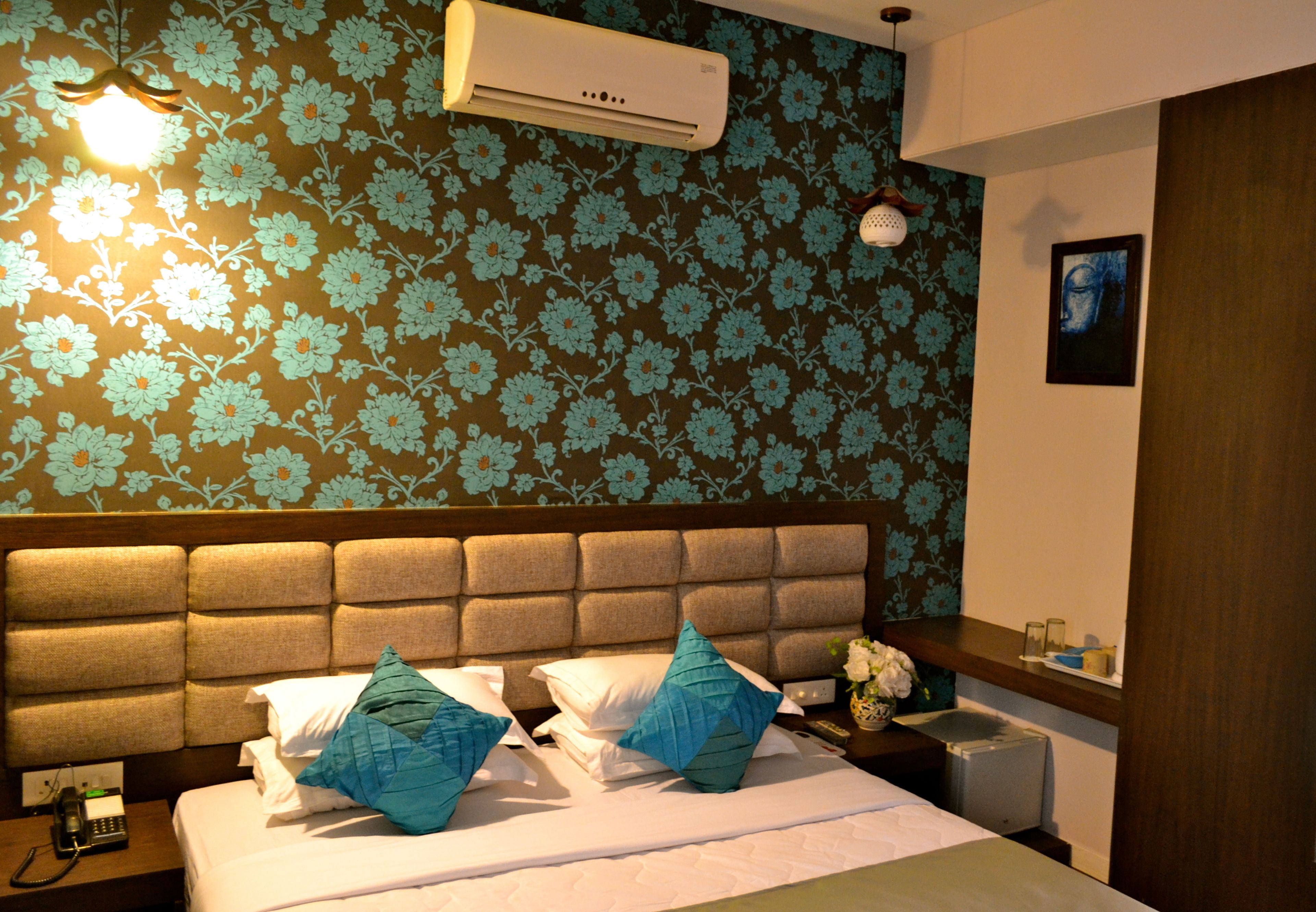 A hotel room with a floral wallpaper, a tufted headboard, a bed with white linens and blue pillows, a bedside table with a phone and a vase of flowers, and an air conditioning unit on the wall.