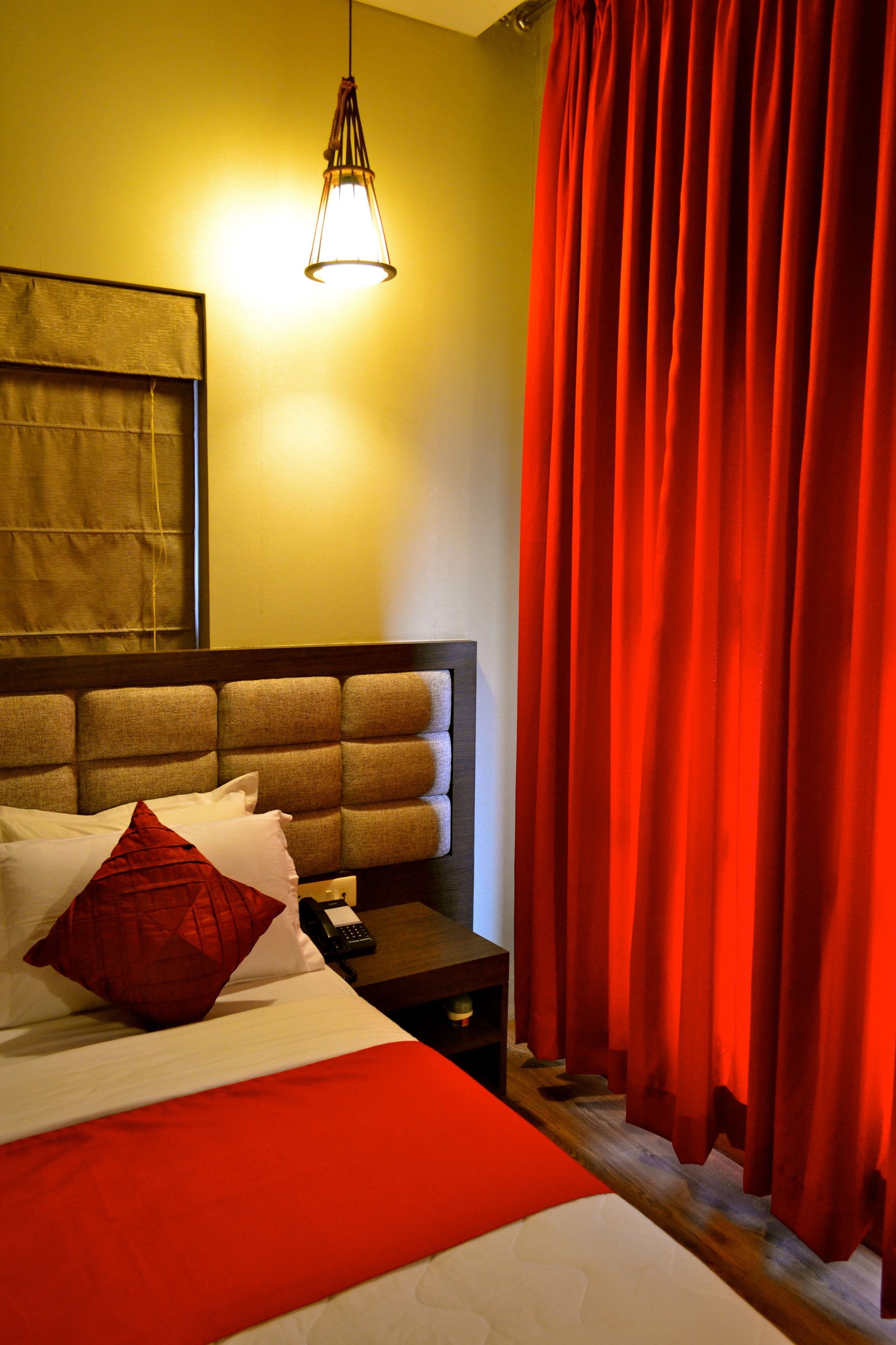 A hotel room with a large bed, a headboard, a bedside table, a lamp, and red curtains.