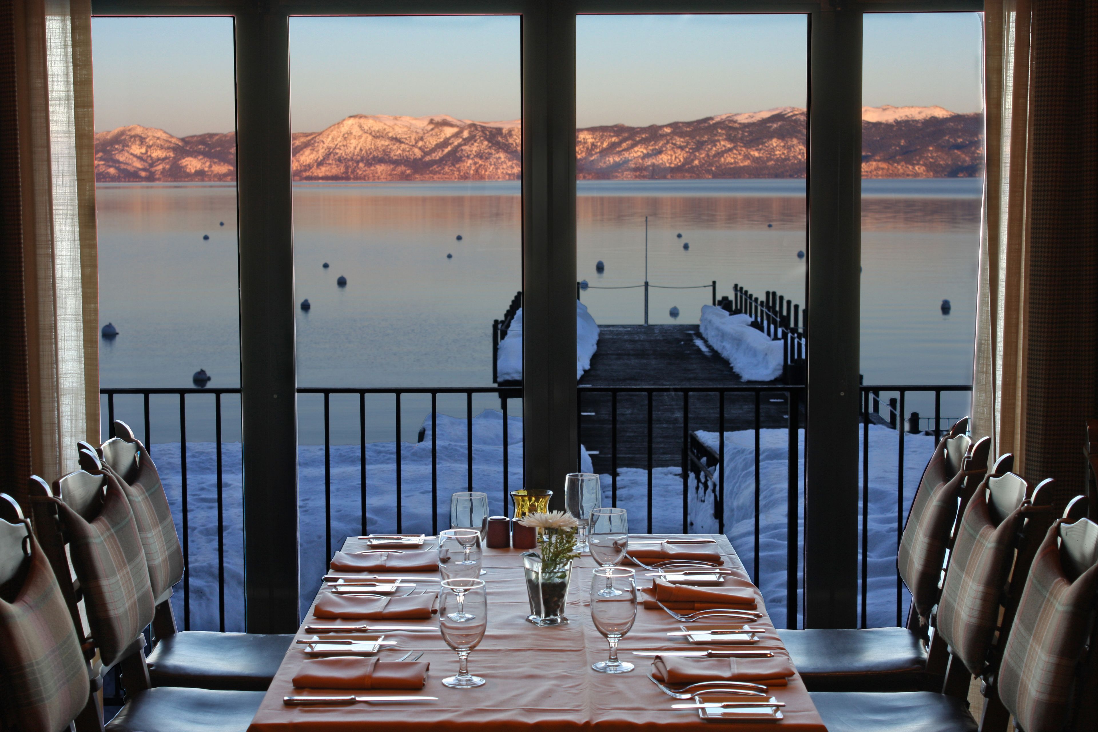 A dining table set for six with a view of a snowy mountain and a dock extending into a calm lake.
