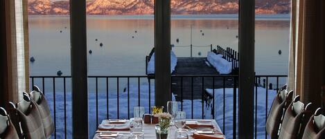 A dining table set for six with a view of a snowy mountain and a dock extending into a calm lake.