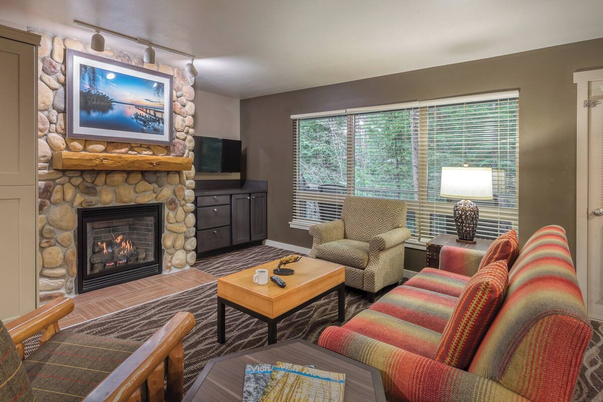 A living room with a stone fireplace, a television mounted above it, a sofa, a coffee table, and a window with blinds.