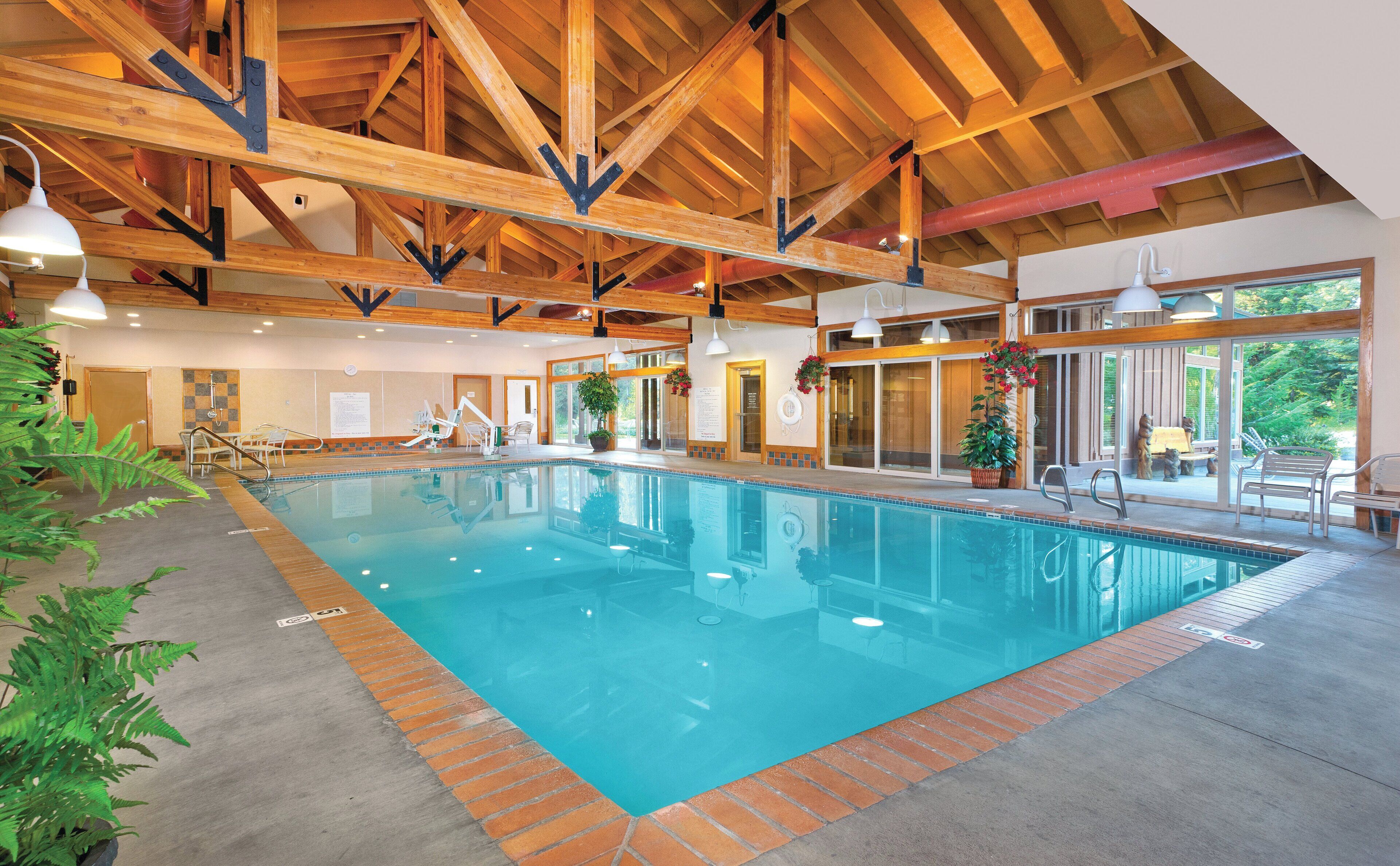 An indoor swimming pool with wooden beams, clear water, and a view of greenery outside.