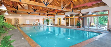 An indoor swimming pool with wooden beams, clear water, and a view of greenery outside.