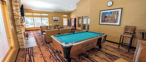 A pool table in a room with a patterned carpet, a clock on the wall, and a window with blinds.