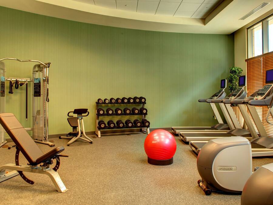 Uma academia com equipamentos de exercícios, um banco, uma bola vermelha de ginástica e um suporte de halteres fixado na parede.