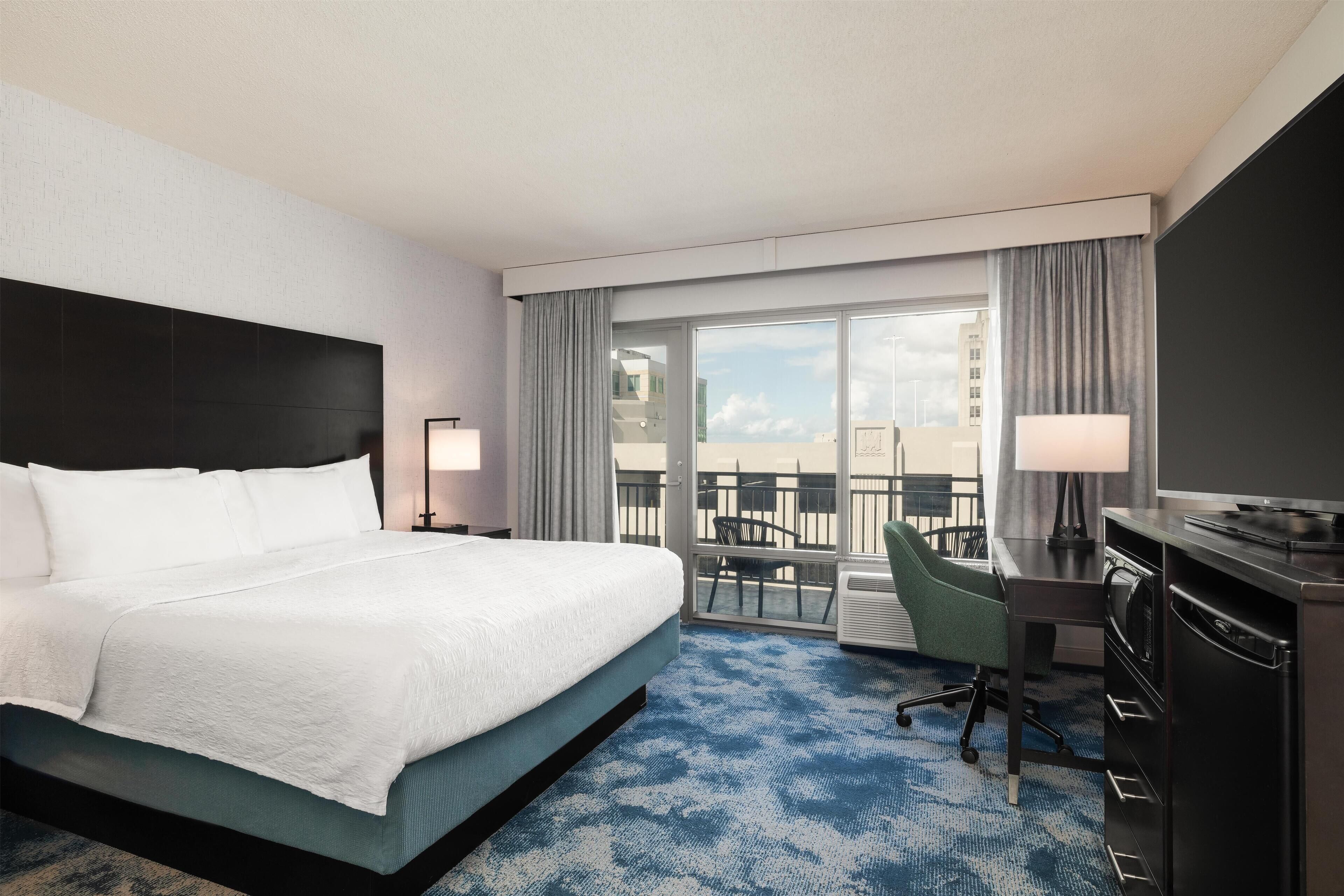 A hotel room with a large bed, a desk with a chair, a TV, and a balcony with a view of buildings.