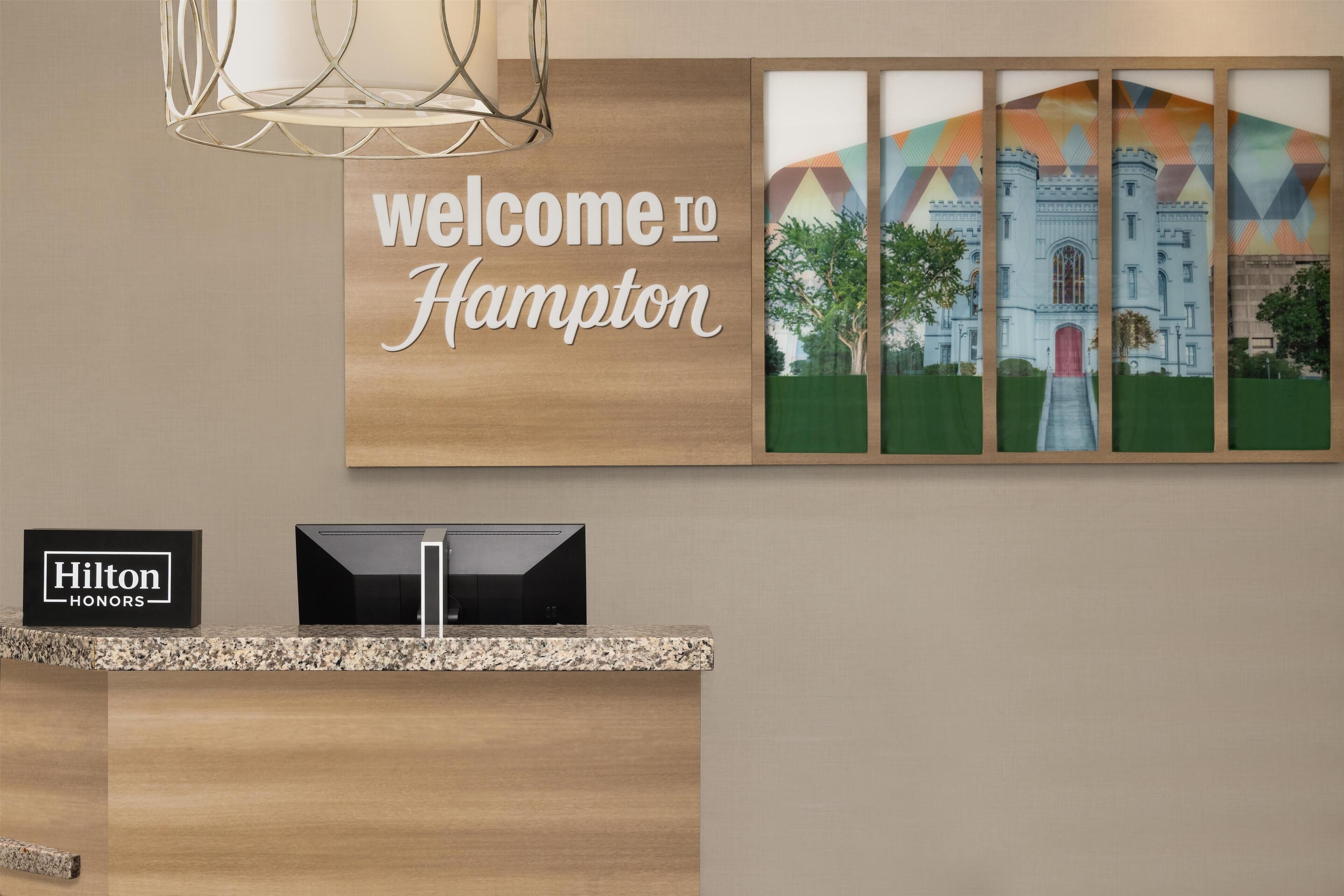Un mostrador de recepción con un letrero que dice "Welcome to Hampton", una fotografía enmarcada de un edificio y un letrero "Hilton Honors".