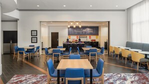 A modern dining area with multiple tables and chairs, a kitchen in the background, and a large window with curtains.