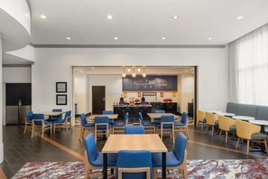 A modern dining area with multiple tables and chairs, a kitchen in the background, and a large window with curtains.