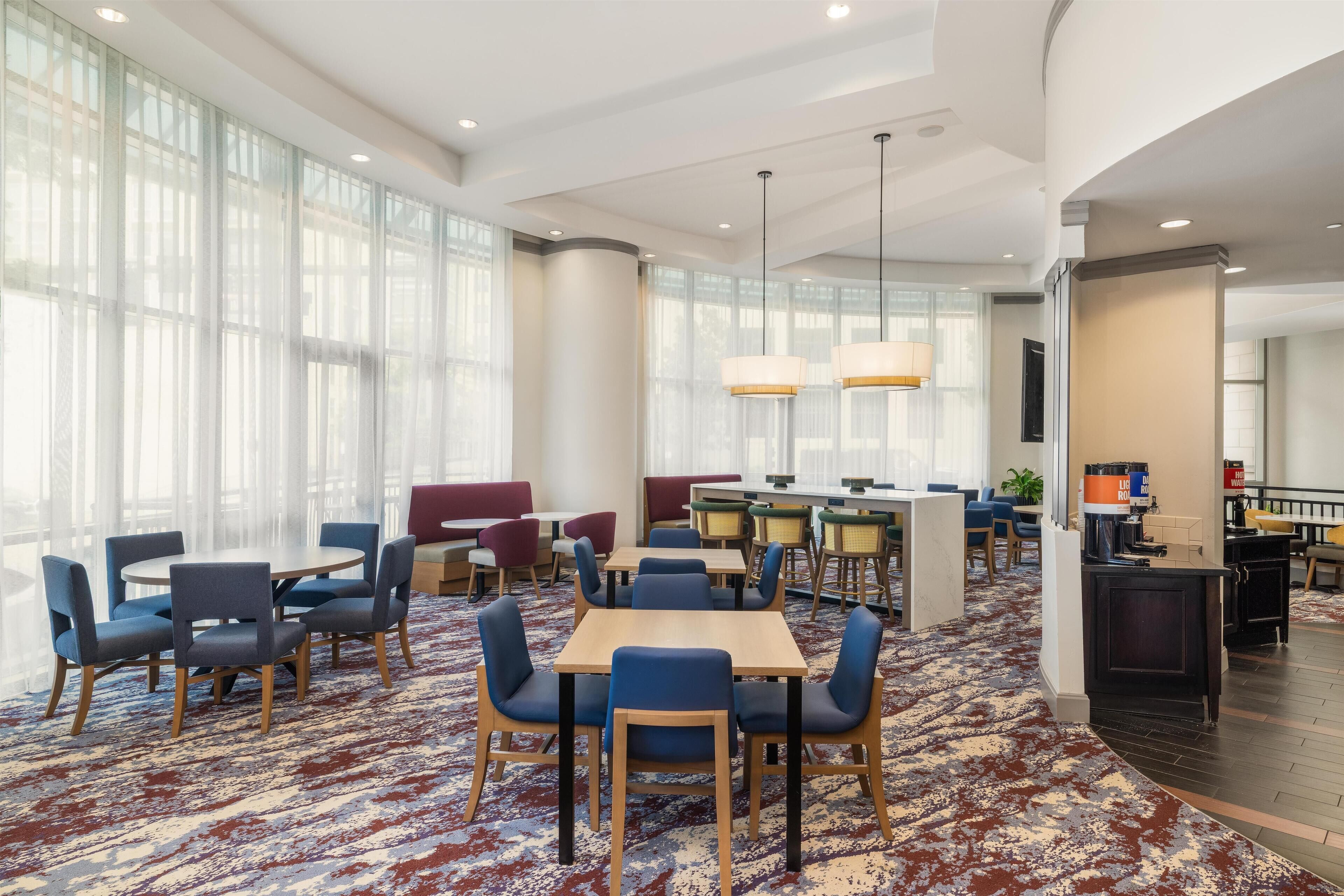 Een ruime hotellobby met ronde tafels en stoelen, een bar en grote ramen die natuurlijk licht binnenlaten.