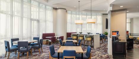 Een ruime hotellobby met ronde tafels en stoelen, een bar en grote ramen die natuurlijk licht binnenlaten.