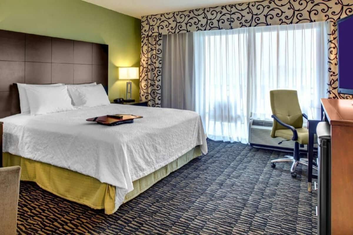 Ein Hotelzimmer mit Bett, Schreibtisch, Bürostuhl, Lampe und einem großen Fenster mit Vorhängen.