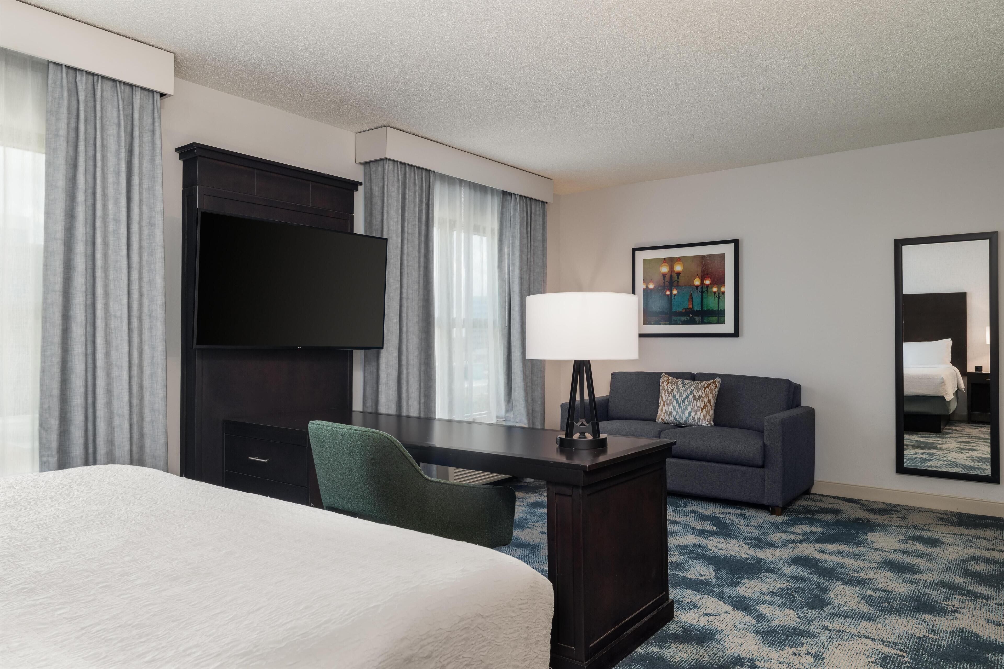 A hotel room with a bed, a desk, a chair, a television, a sofa, a lamp, a mirror, and a painting on the wall.