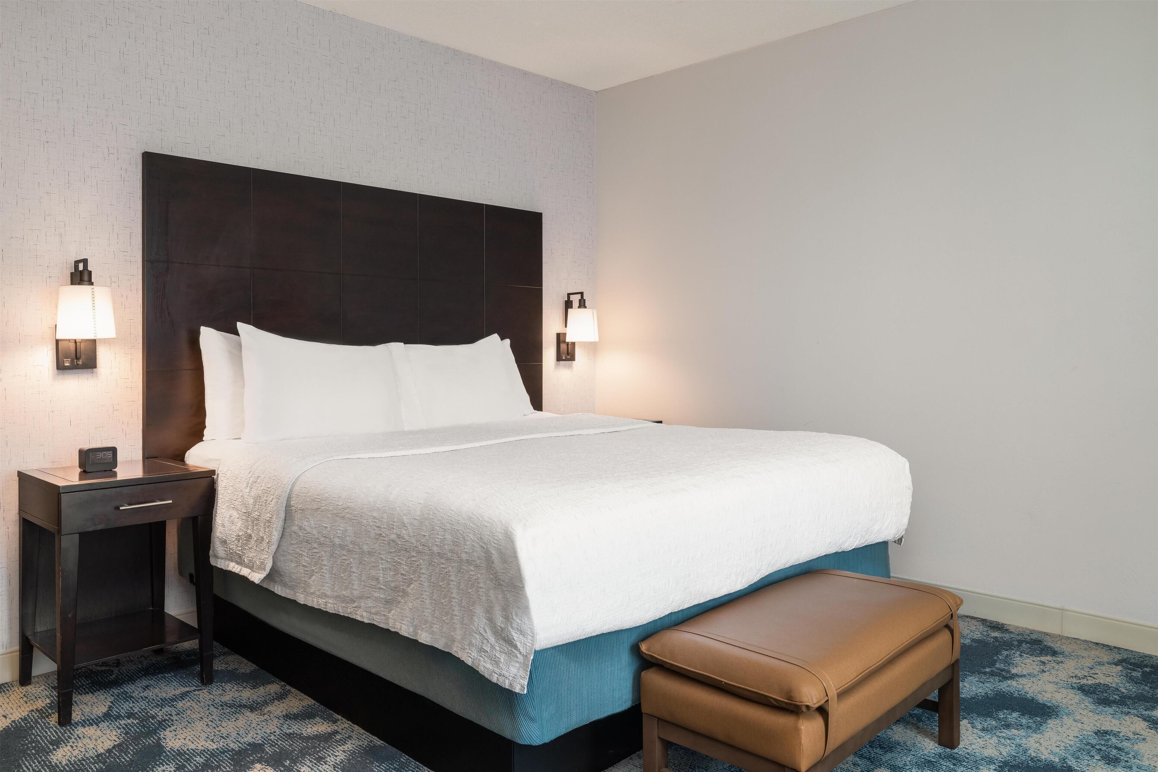 A hotel room with a large bed, two bedside tables, a nightstand, a bench, and two wall-mounted lamps.