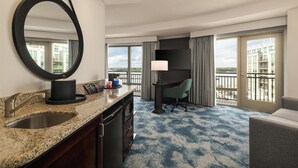 Une chambre d’hôtel dotée d’un grand miroir, d’un plan de travail en granit avec un évier, d’un bureau avec une chaise et offrant une vue sur la ville grâce à de grandes fenêtres.