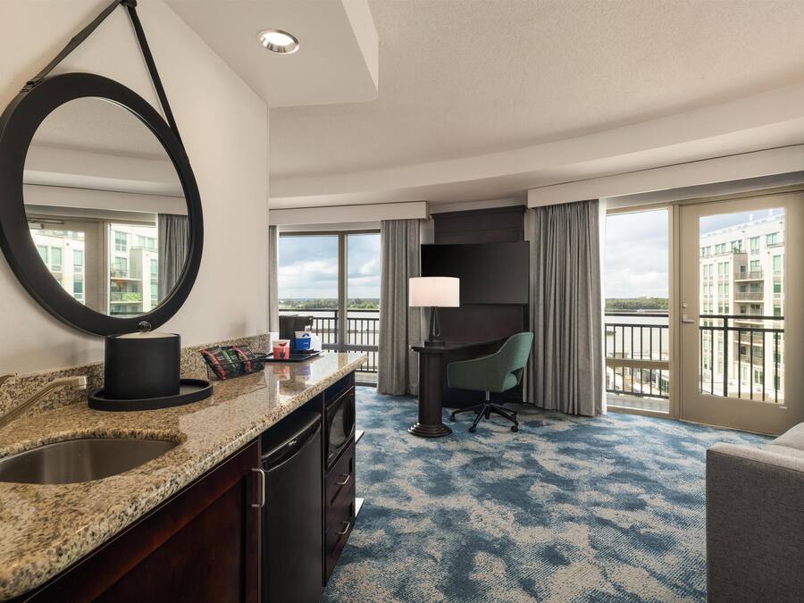 A hotel room with a large mirror, a granite countertop with a sink, a desk with a chair, and a view of the city through large windows.
