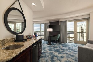 Habitación de hotel con un espejo grande, encimera de granito con lavamanos, escritorio con silla y vistas a la ciudad a través de amplias ventanas.