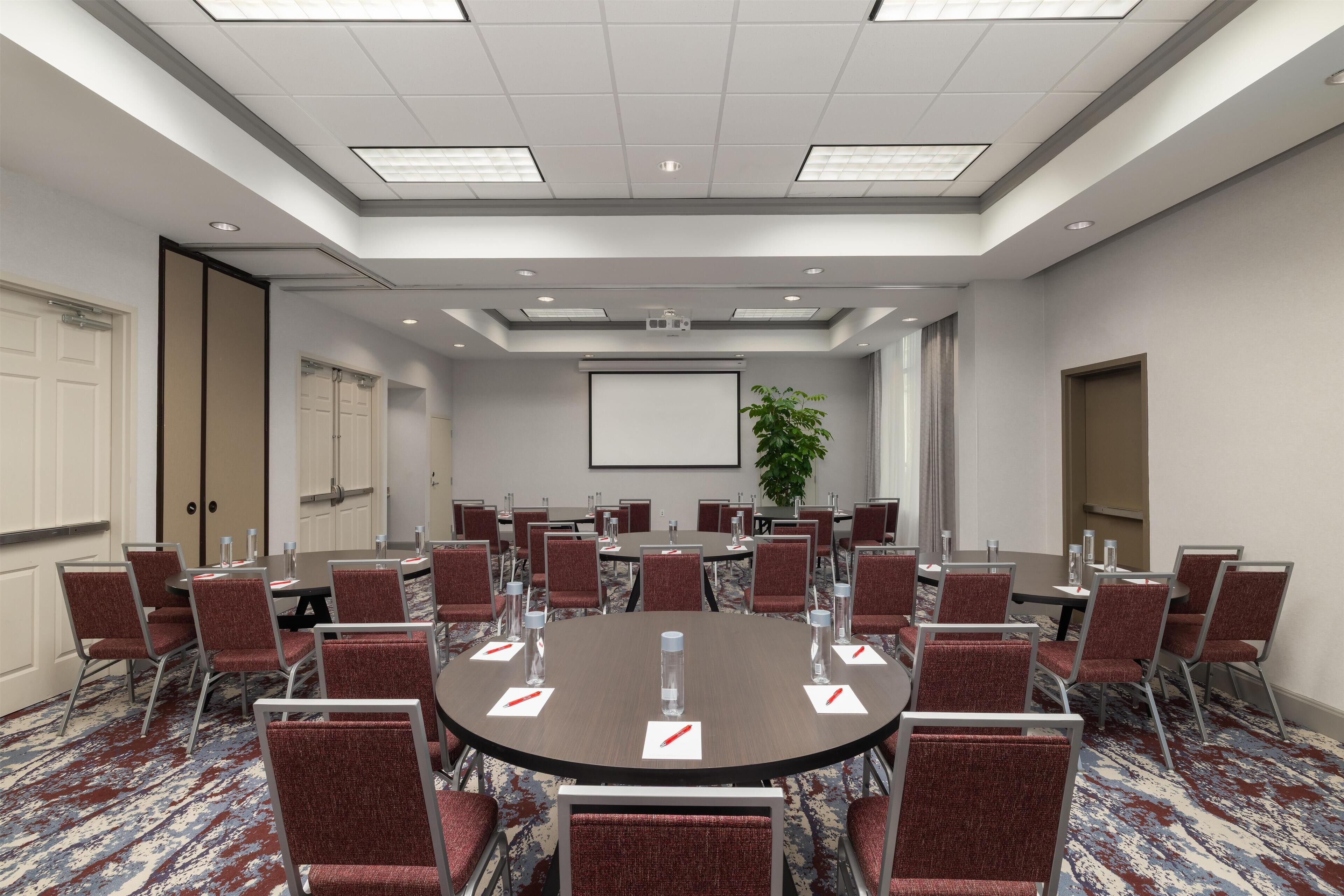 Uma sala de conferências com uma grande mesa redonda, cadeiras vermelhas e uma tela de projetor.