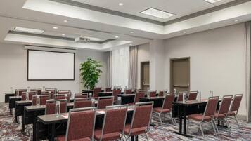 Un salón de conferencias con varias mesas y sillas, una pantalla de proyector y una planta grande.