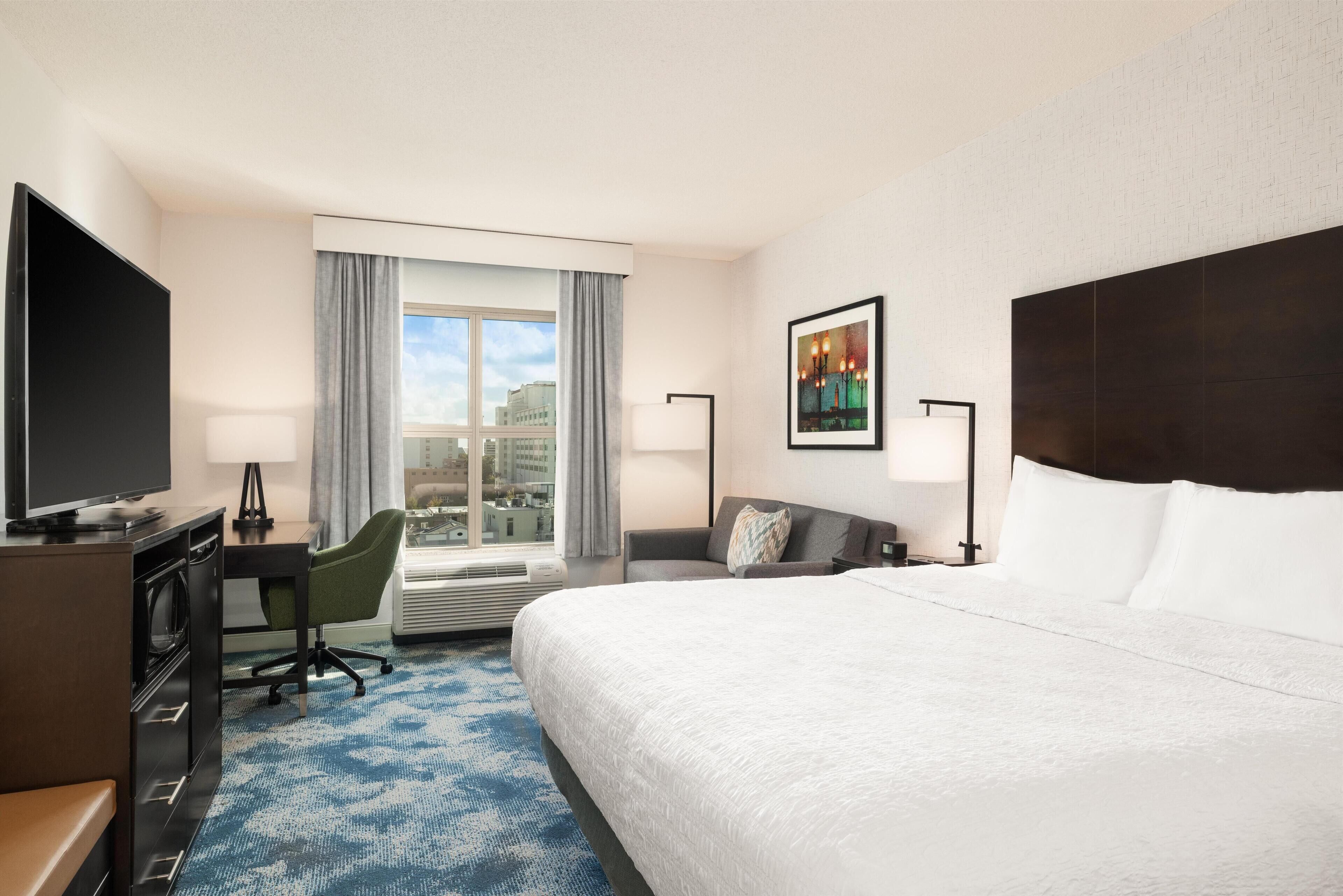 A hotel room with a large bed, a desk with a computer, a TV, a chair, a window with curtains, and a city view.