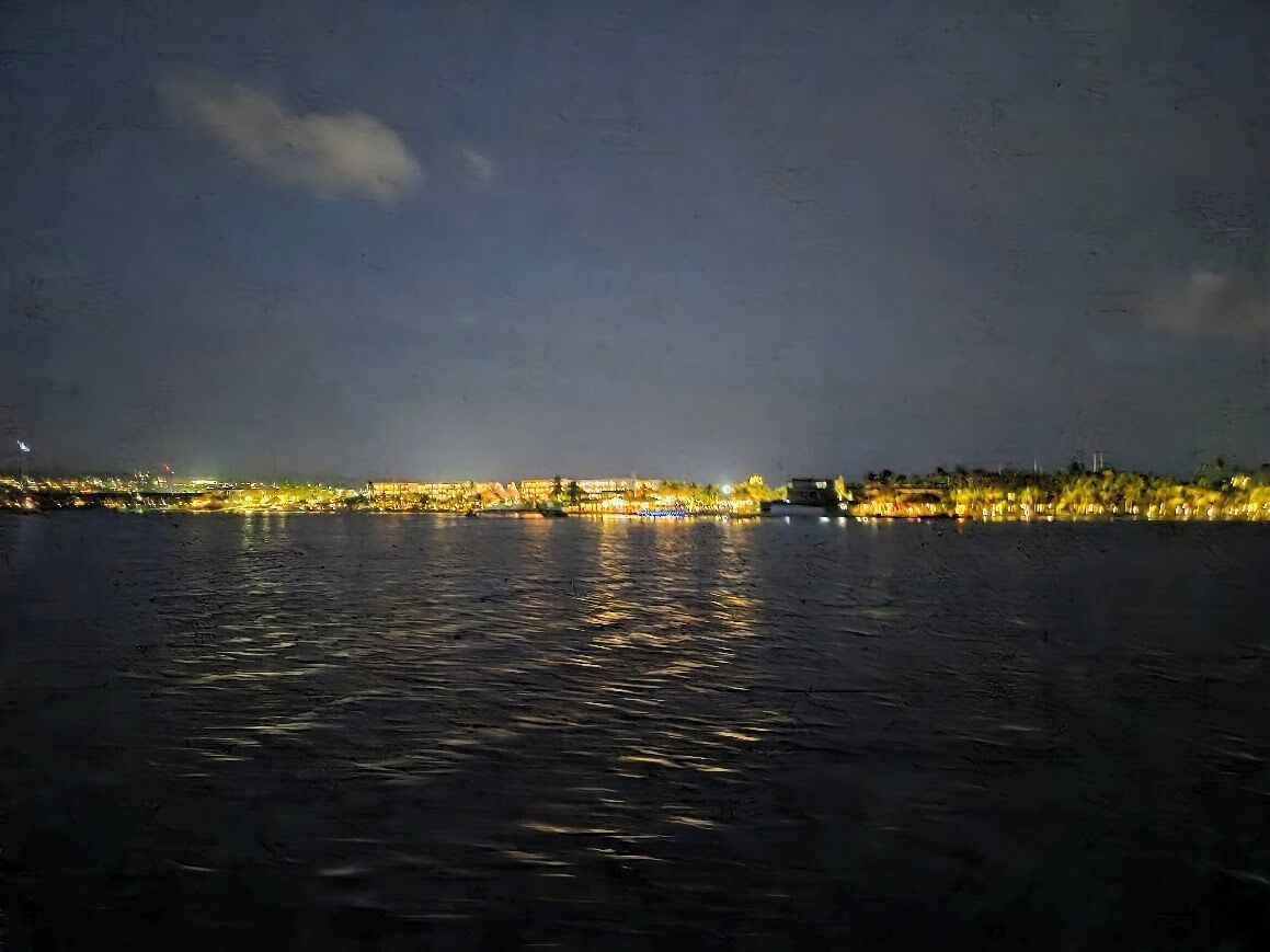 Vista nocturna de un paisaje urbano frente al agua, con edificios iluminados que se reflejan en la superficie.