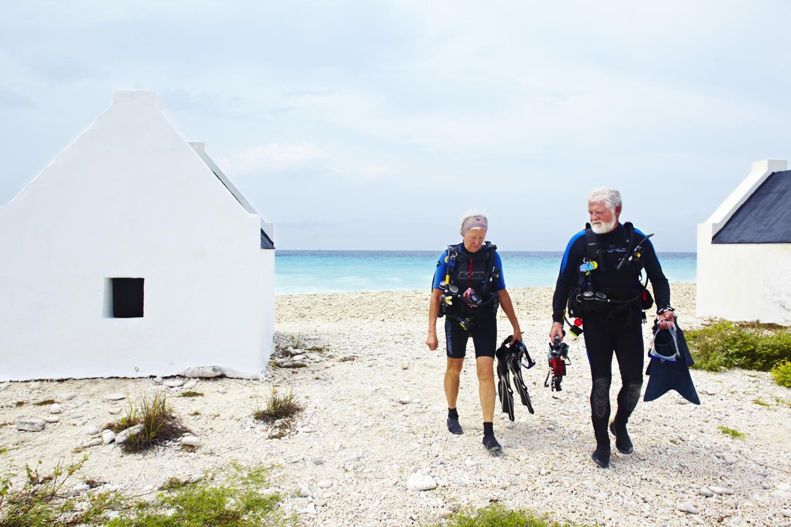 Deux personnes se promènent sur une plage, avec des bâtiments blancs en arrière-plan.