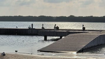 Ein Uferbereich mit einer Holzstege, der sich in ruhiges Wasser erstreckt, umgeben von einem Sandstrand und einer in der Ferne liegenden, bewaldeten Küste.