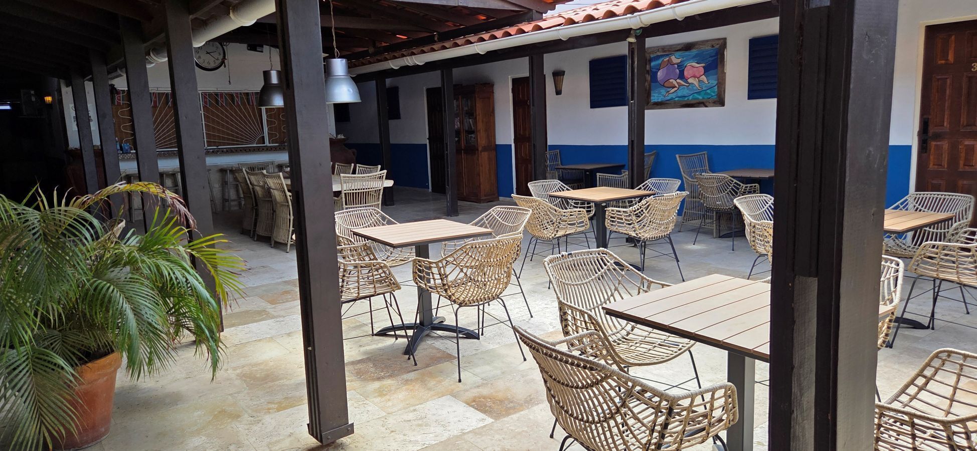 An outdoor café area with wicker tables and chairs, a potted plant, and a clock on the wall.