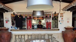 A rustic outdoor dining area with a large table and wicker chairs, a hanging light fixture, and a bar in the background. - The Lodge Bonaire (Kralendijk)