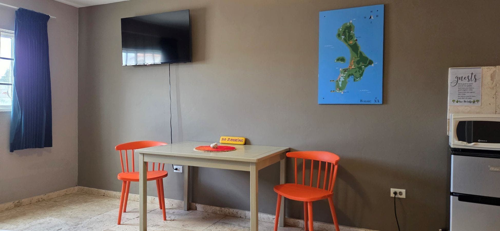 Ein Zimmer mit an der Wand befestigtem Fernseher, einer Karte, einem Tisch mit Stühlen und einem Verbotsschild für Rauchen.