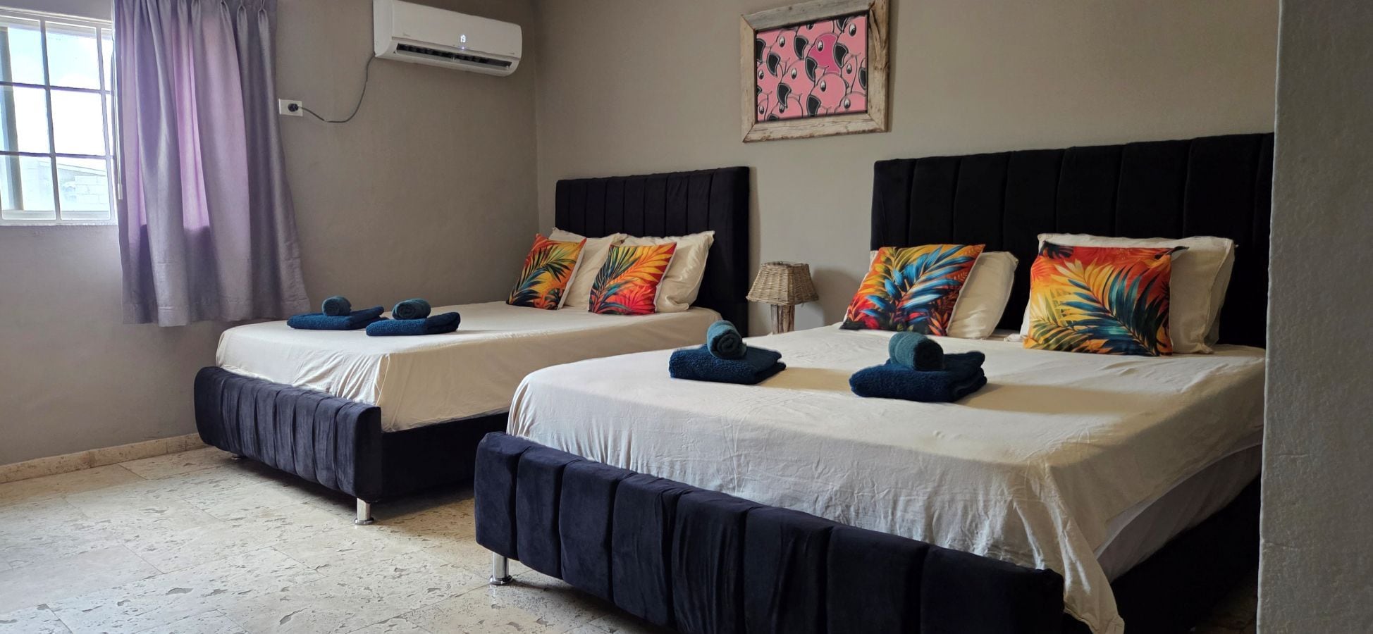 Two twin beds with matching headboards and footboards, a window with curtains, an air conditioning unit, and a wall-mounted artwork.