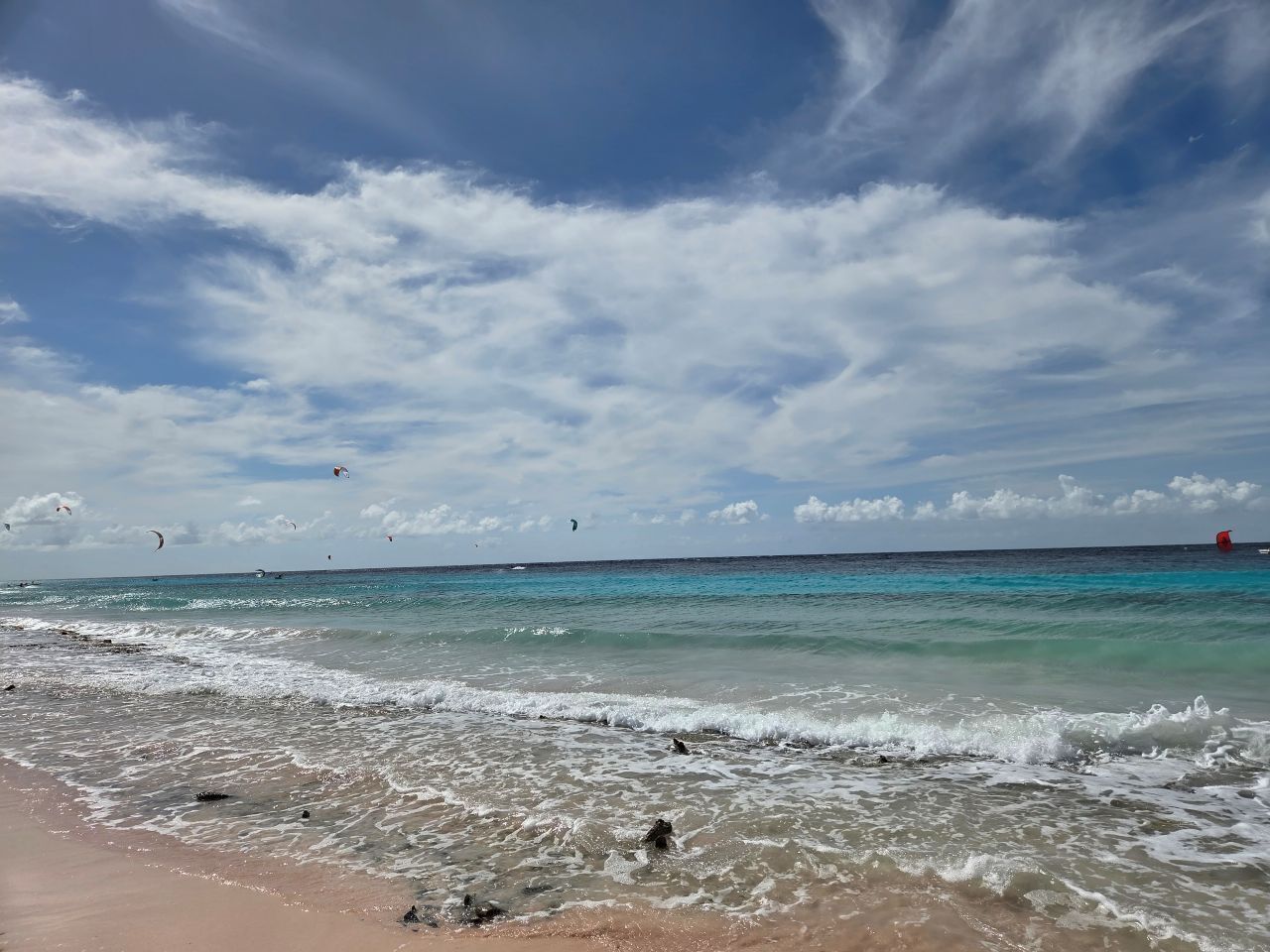 Una playa con cielos despejados, aguas tranquilas y cometas a lo lejos.