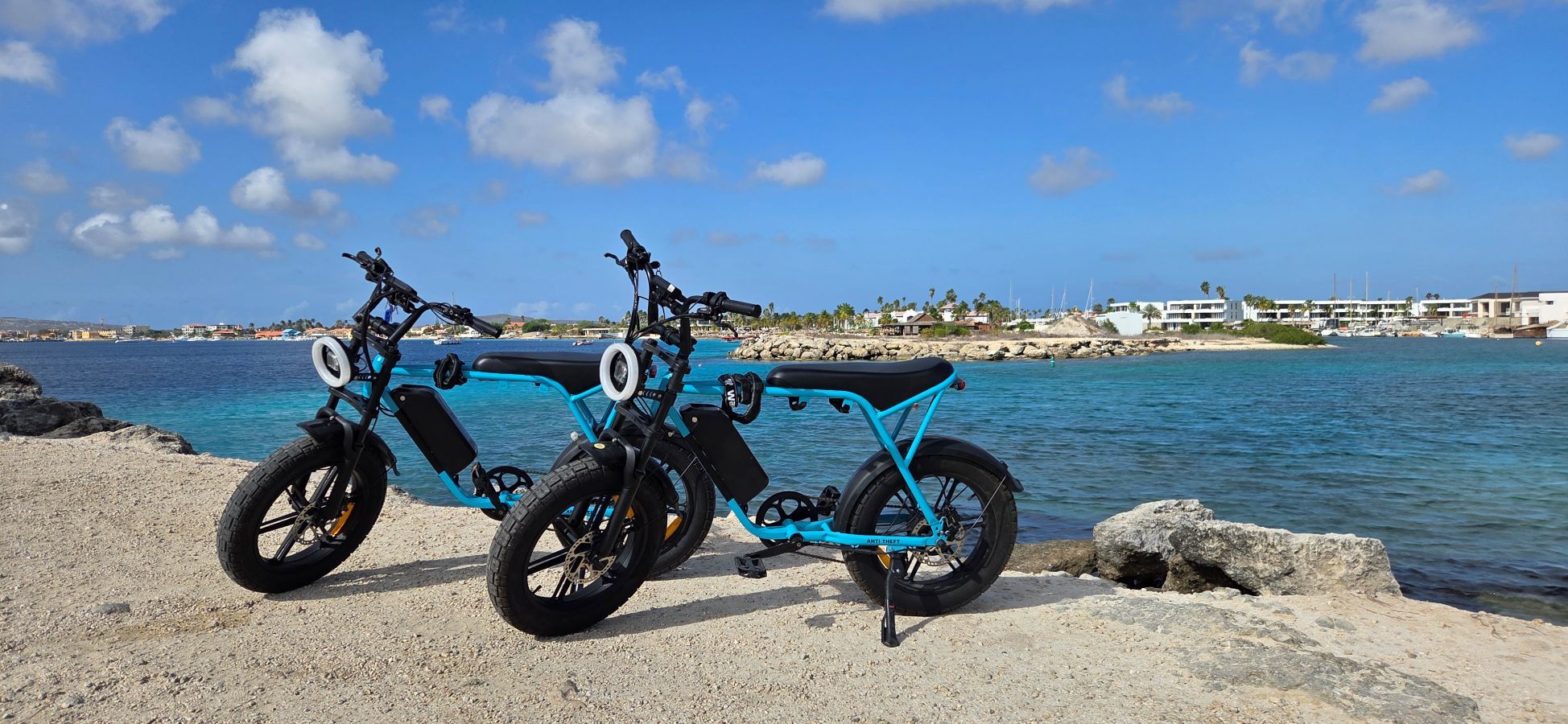Dos bicicletas eléctricas estacionadas en una costa rocosa con un cielo azul despejado y un mar tranquilo al fondo.