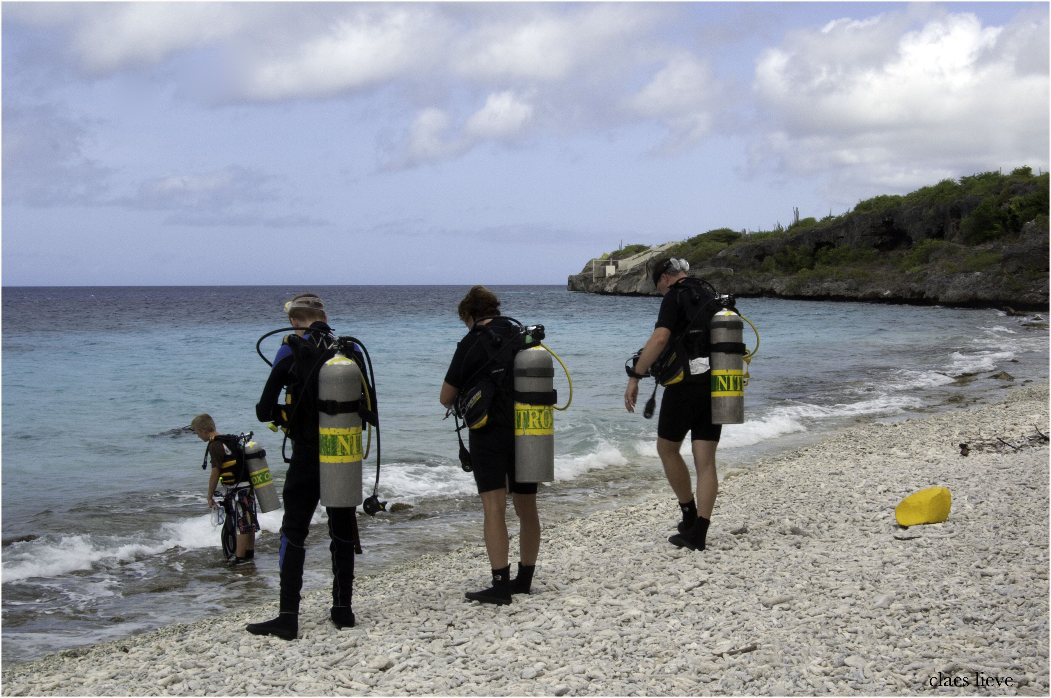 A group of scuba divers on a pebbly beach with clear blue water and a rocky outcrop in the background.
