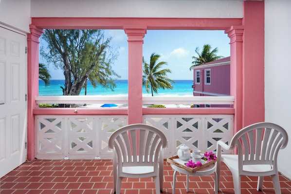 Un balcón con dos sillas blancas, una bandeja con tetera, tazas y flores, con vistas a una playa con palmeras y un edificio.