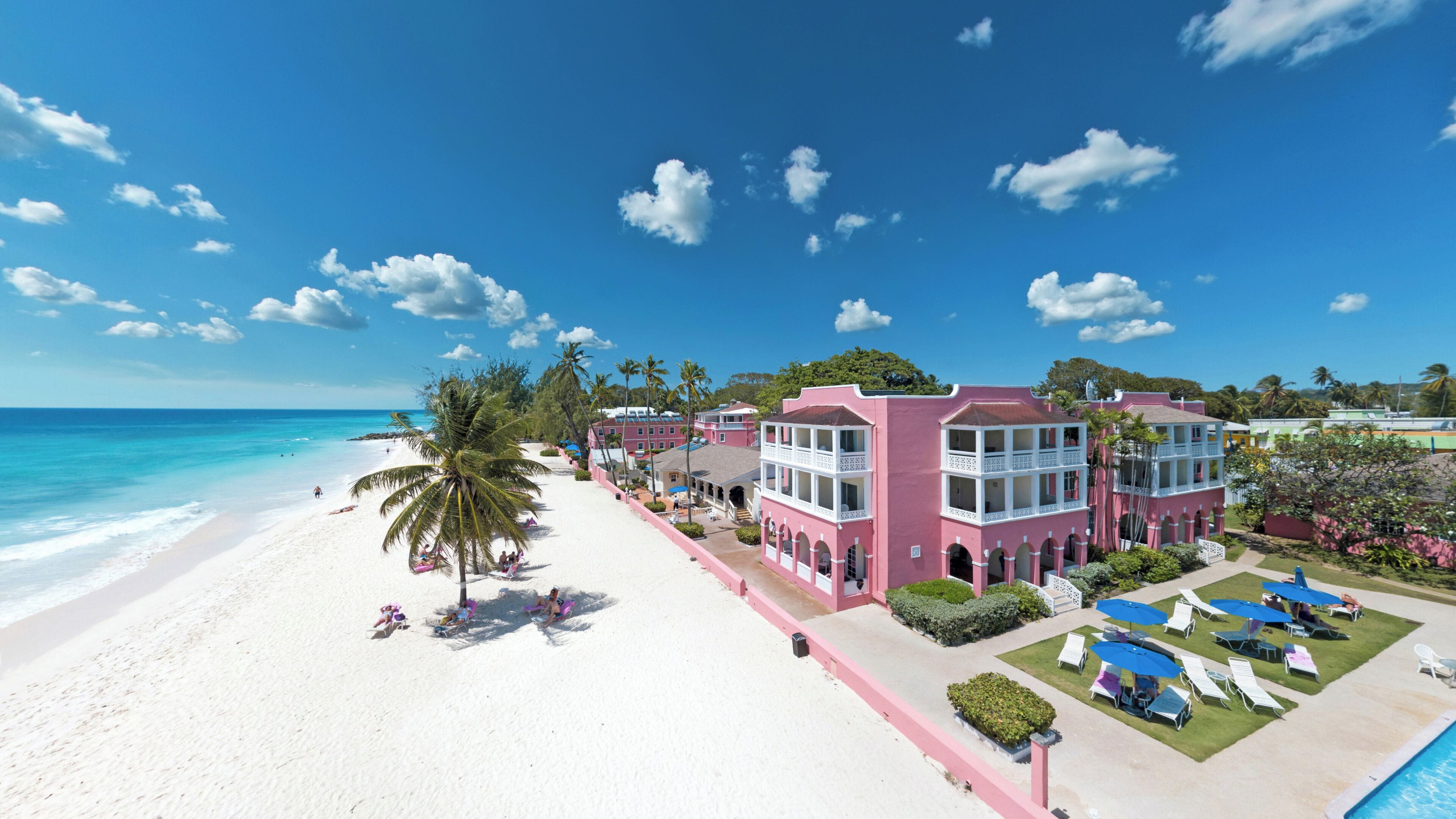 A beachfront hotel with a white sandy beach, palm trees, and a clear blue sky.