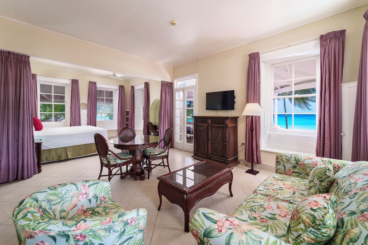 A hotel room with a bed, a dining table with chairs, a sofa, a TV, and a view of the ocean.